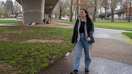 a person walks in a park with grass and a road bridge above her