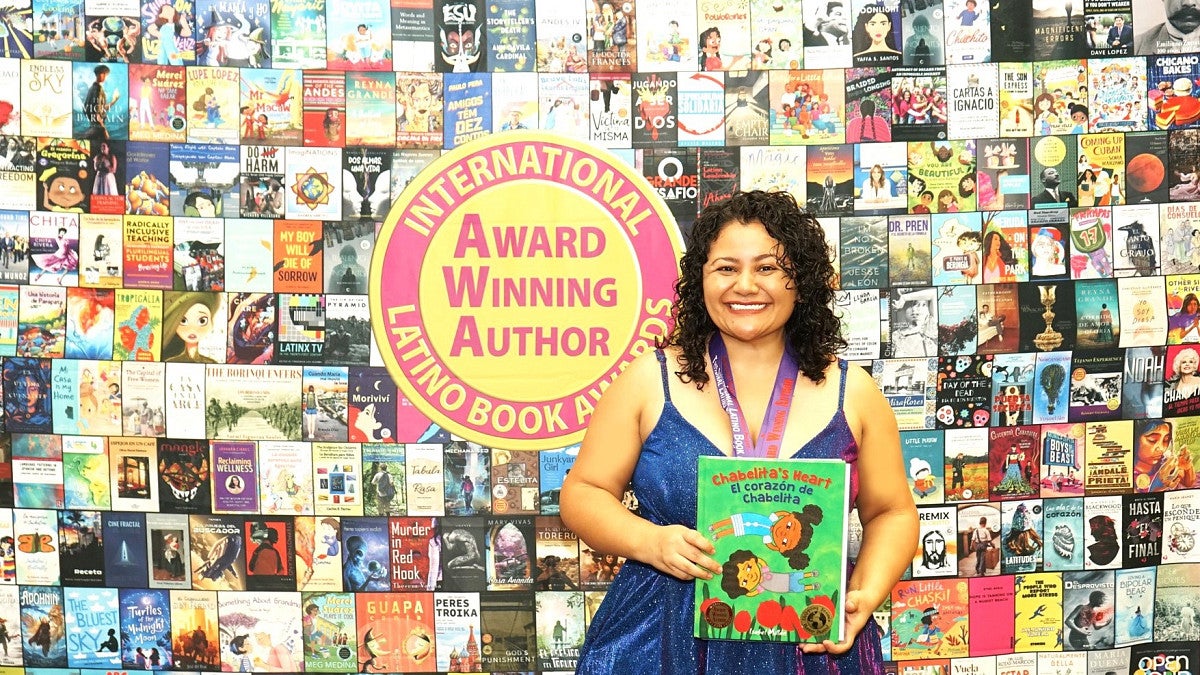 Isabel Millán stands in front of a poster of book covers holding her book