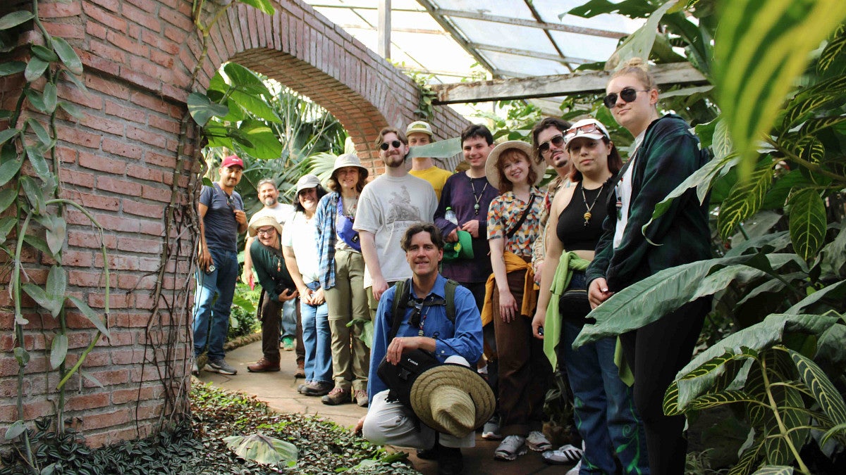 a group of people pose outside of a brick building surrounded by lush green plants