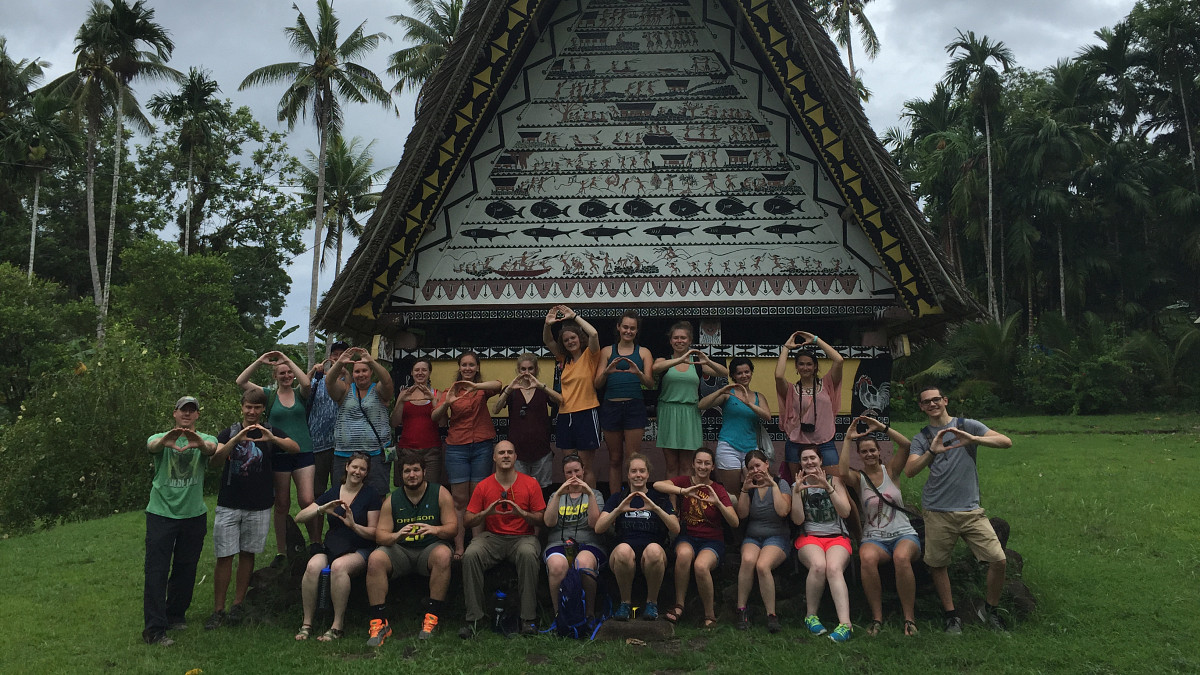 group of students throwing "O" symbol with hands in front of small house with palm trees in background